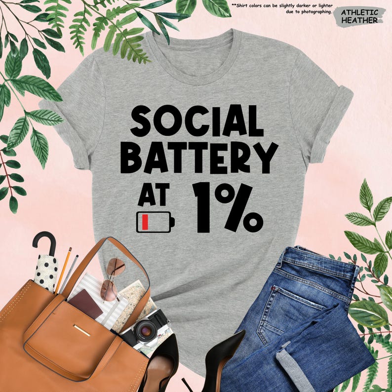 May include: Athletic heather gray t-shirt with the text "SOCIAL BATTERY AT 1%" in black. The design includes a low battery icon. The shirt is paired with blue jeans, a brown purse, and black heels.