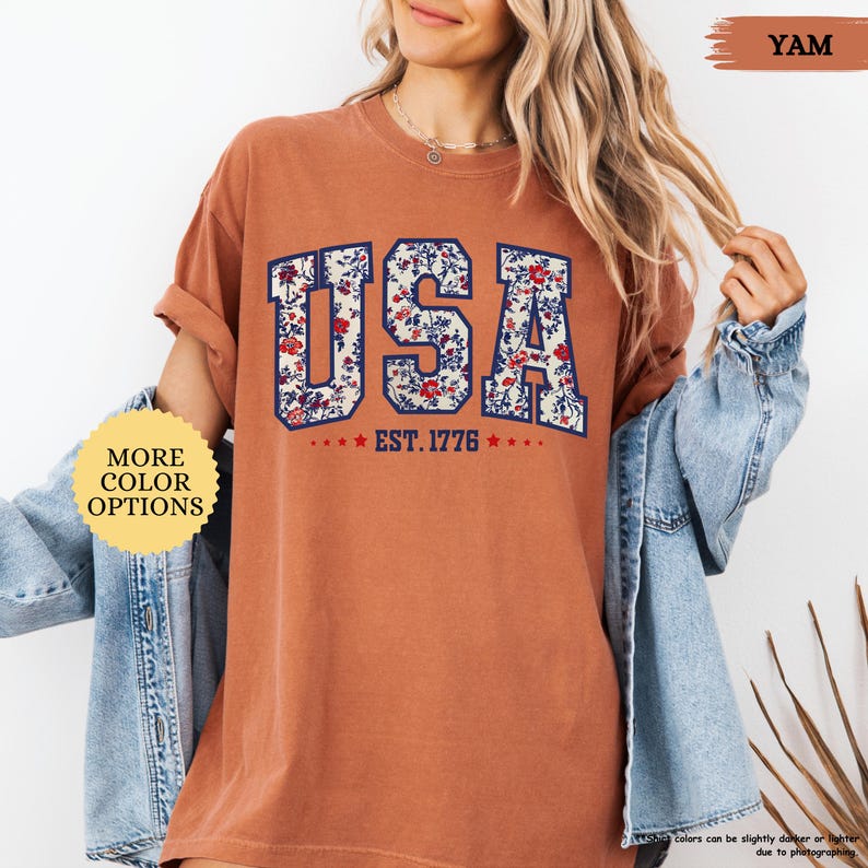 May include: A burnt orange t-shirt with a floral print design of the word "USA" and the text "EST. 1776" in navy blue and white.