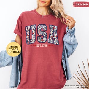 May include: A red t-shirt with a floral print design of the word "USA" and the text "EST. 1776".