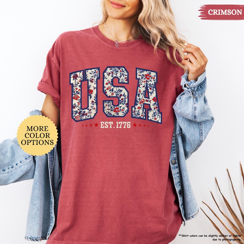 May include: A red t-shirt with a floral print design of the word "USA" and the text "EST. 1776".