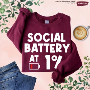 May include: Maroon sweatshirt with the text "SOCIAL BATTERY AT 1%" in white. A low battery icon is also displayed. The sweatshirt is paired with blue jeans and a cup of coffee with latte art.