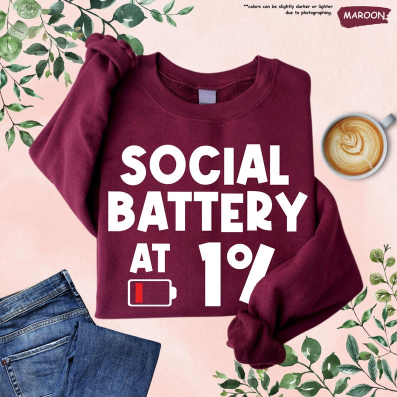 May include: Maroon sweatshirt with the text "SOCIAL BATTERY AT 1%" in white. A low battery icon is also displayed. The sweatshirt is paired with blue jeans and a cup of coffee with latte art.