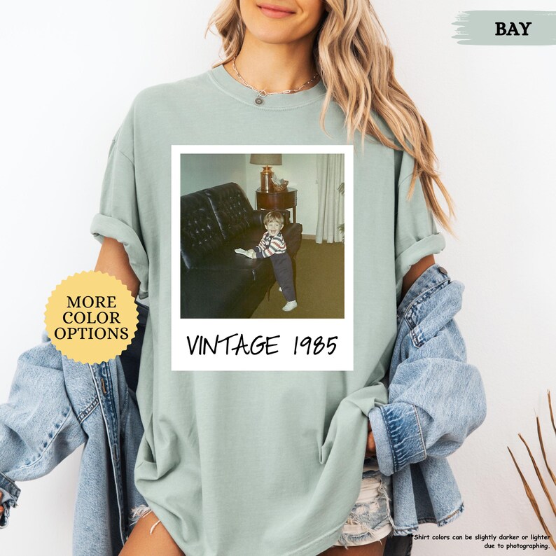Op de afbeelding: Een lichtgroen T-shirt met een vintage Polaroid-foto van een jonge jongen die op een bank zit. De foto is omlijst met een witte rand en de tekst "Vintage 1985" is onder de foto gedrukt.