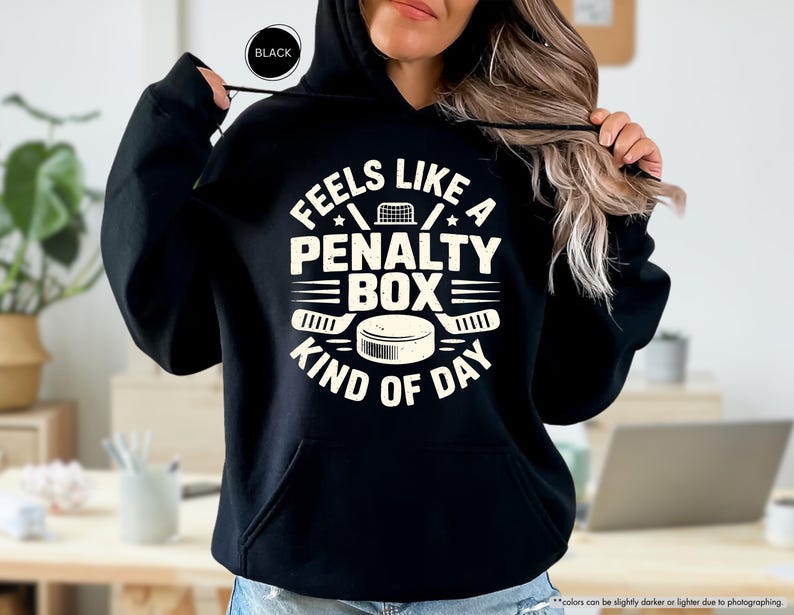 May include: Black hoodie with the text "FEELS LIKE A PENALTY BOX KIND OF DAY" in white. The design includes hockey sticks, a hockey puck, and a hockey net. The hoodie has a hood and a front pocket.