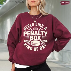 May include: Maroon sweatshirt with the text "FEELS LIKE A PENALTY BOX KIND OF DAY" in white. The design includes hockey sticks, a hockey net, and a hockey puck. The sweatshirt is a long-sleeved crew neck.