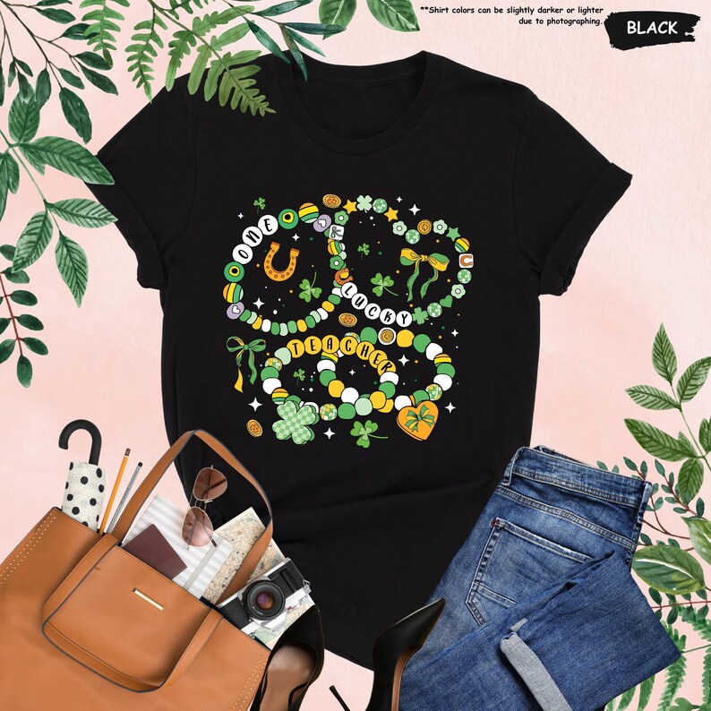 K&ouml;nnte beinhalten: Schwarzes T-Shirt mit einem gr&uuml;nen, gelben und wei&szlig;en Kleeblatt-Design. Das Kleeblatt besteht aus Perlen und tr&auml;gt die Aufschrift "One Lucky Teacher".