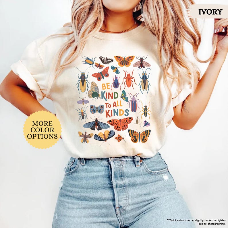 May include: Ivory t-shirt with a colourful insect and butterfly graphic and the text "BE KIND TO ALL KINDS." The shirt has a crew neck and short sleeves. Additional text on the shirt says "MORE COLOR OPTIONS."
