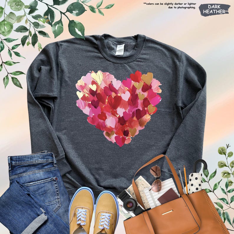 Pu&ograve; includere: Felpa grigio scuro con un design a cuore composto da pennellate rosse, rosa e oro. L'immagine include anche jeans blu, scarpe color camoscio, una borsa tote marrone e accessori.