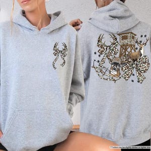 May include: Sport grey hoodie with a front pocket and a hood. The front features a bow with deer antlers. The back of the hoodie has a deer, bow, hunting stand, and footprints design. The bow and deer designs have a camouflage pattern.
