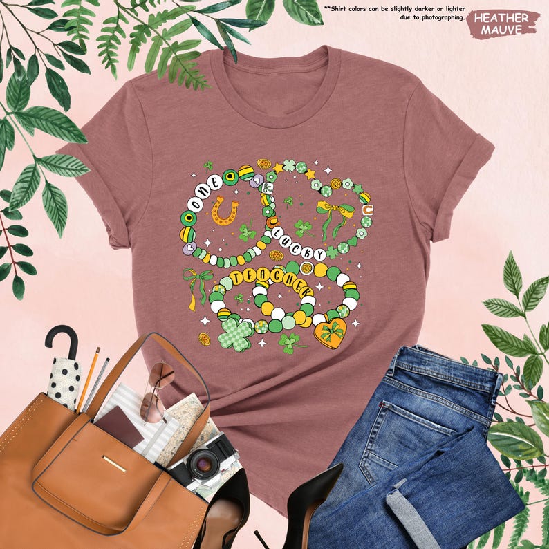 K&ouml;nnte beinhalten: Heather-mauvefarbenes T-Shirt mit einem Motiv aus drei gr&uuml;nen, wei&szlig;en und goldenen Perlenarmb&auml;ndern mit den Worten "One Lucky Teacher" darauf. Die Armb&auml;nder sind mit Kleebl&auml;ttern, Hufeisen und Schleifen verziert.