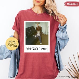 Op de afbeelding: Een crimson t-shirt met een vintage Polaroid-foto van een jong kind dat op een bank zit. De foto is omlijst met een witte rand en de tekst "Vintage 1985" is onder de foto gedrukt.