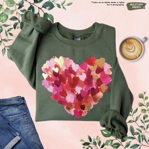 Pu&ograve; includere: Felpa verde militare con un design a cuore composto da cuori a pennellate in rosso, rosa e oro. La felpa ha un girocollo e maniche lunghe. Il testo "colors can be slightly darker or lighter due to photographing" &egrave; visibile.
