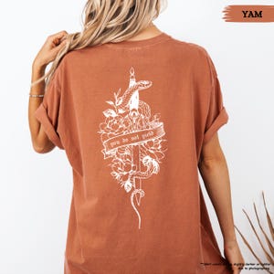 May include: A rust-coloured t-shirt with a white graphic on the back. The graphic shows a snake coiled around a sword, flowers, and the text "you do not yield" on a banner. The shirt has rolled-up sleeves.
