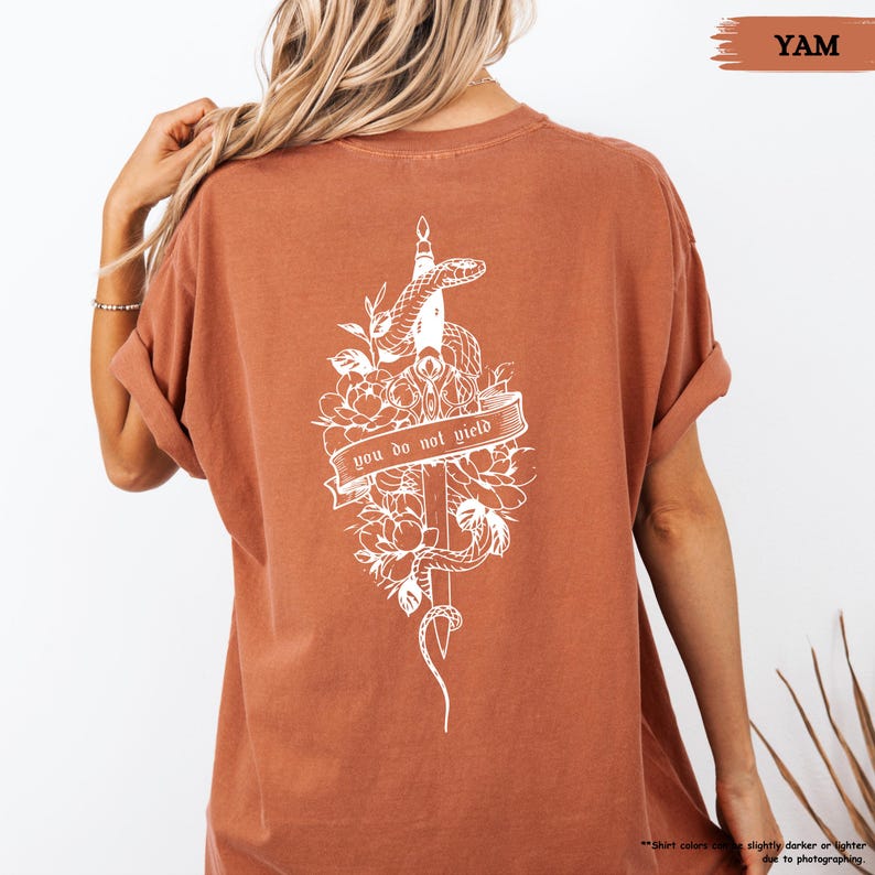 May include: A rust-coloured t-shirt with a white graphic on the back. The graphic shows a snake coiled around a sword, flowers, and the text "you do not yield" on a banner. The shirt has rolled-up sleeves.