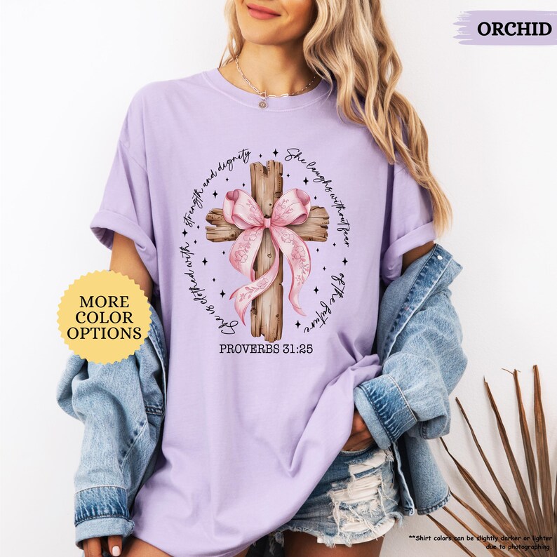 Op de afbeelding: Een lichtpaarse t-shirt met een houten kruis en een roze lint. Het kruis is omgeven door de tekst "Zij lacht zonder angst voor de toekomst" en "gekleed met kracht en waardigheid". De tekst "Spreuken 31:25" staat onder het kruis.