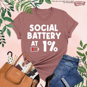 May include: Heather mauve t-shirt with the text "SOCIAL BATTERY AT 1%" in white. The graphic includes a low battery icon. The shirt is paired with blue jeans, a brown purse, and black heels.