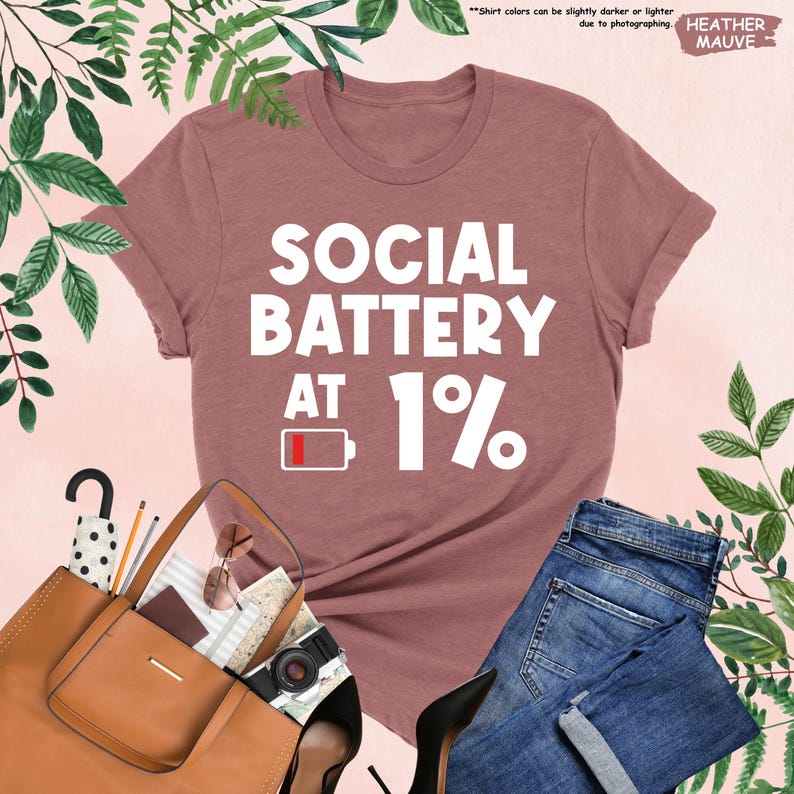 May include: Heather mauve t-shirt with the text "SOCIAL BATTERY AT 1%" in white. The graphic includes a low battery icon. The shirt is paired with blue jeans, a brown purse, and black heels.
