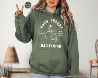 May include: A green hooded sweatshirt with the text "Hard Fought" in white lettering. The text is surrounded by a circle with boxing gloves inside. The text "Heartfelt" and "Been Through Hell" are below the circle. The text "Hallelujah" is below the boxing gloves.