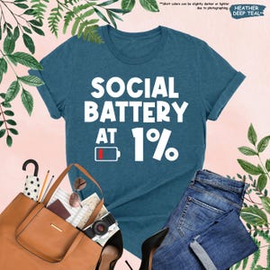 May include: A heather deep teal t-shirt with the text "SOCIAL BATTERY AT 1%" in white. The design includes a low battery icon. The shirt is paired with blue jeans, a brown tote bag, and black heels.