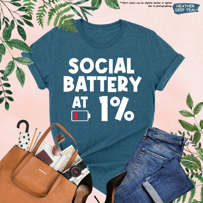 May include: A heather deep teal t-shirt with the text "SOCIAL BATTERY AT 1%" in white. The design includes a low battery icon. The shirt is paired with blue jeans, a brown tote bag, and black heels.