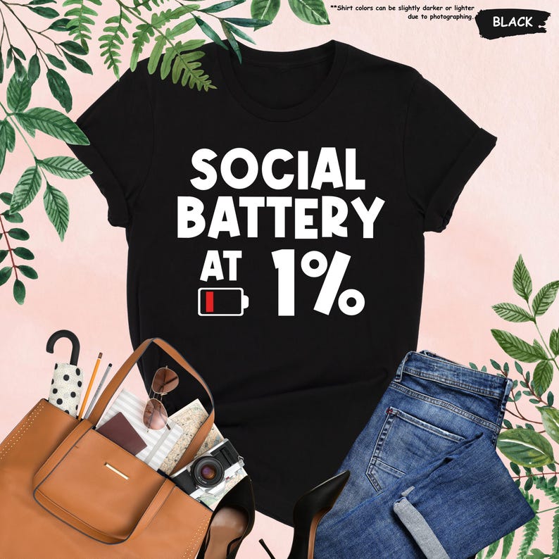 May include: Black t-shirt with the text "SOCIAL BATTERY AT 1%" in white. The design includes a battery icon at 1%. The shirt is displayed with a brown tote bag, sunglasses, a camera, and blue jeans.