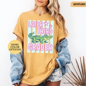 May include: Mustard-colored t-shirt with the text "Later Third Grader" in pink and a cartoon alligator. The shirt also has a light blue and white striped background. The words "More Color Options" are also visible.