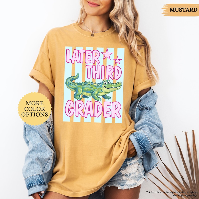 May include: Mustard-colored t-shirt with the text "Later Third Grader" in pink and a cartoon alligator. The shirt also has a light blue and white striped background. The words "More Color Options" are also visible.