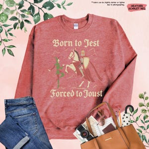 K&ouml;nnte beinhalten: Heather-scharlachrotes Sweatshirt mit dem Text "Born to Jest Forced to Joust" und einer Grafik eines Narren und eines Ritters zu Pferd. Das Sweatshirt hat einen Rundhalsausschnitt.