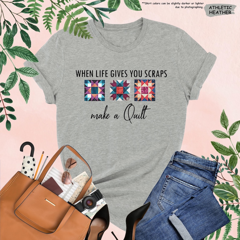 May include: Athletic heather gray t-shirt with the text "WHEN LIFE GIVES YOU SCRAPS make a Quilt." The shirt features three colorful quilt block designs. A brown leather tote bag, blue jeans, and black heels are also visible.