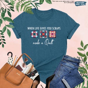 May include: A teal t-shirt with the text "WHEN LIFE GIVES YOU SCRAPS make a Quilt" above three colorful quilt block designs. The shirt is displayed with a brown tote bag, sunglasses, a camera, and blue jeans.