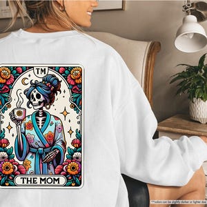 May include: White sweatshirt with a colorful graphic of a skeleton in a robe holding a cup of coffee. The graphic is surrounded by flowers and the text "TM THE MOM".