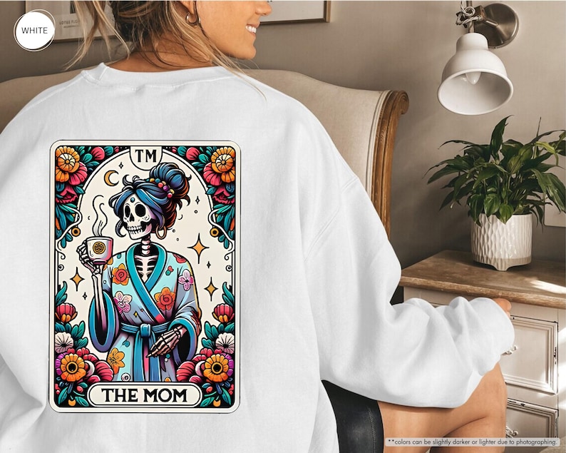May include: White sweatshirt with a colorful graphic of a skeleton in a robe holding a cup of coffee. The graphic is surrounded by flowers and the text "TM THE MOM".