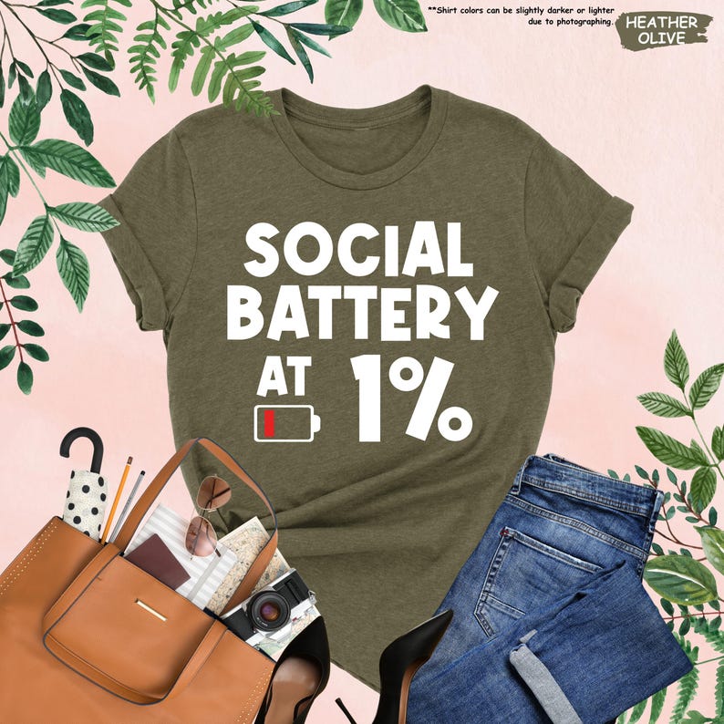 May include: Heather olive green t-shirt with the text "SOCIAL BATTERY AT 1%" in white. The design includes a low battery symbol. The shirt is paired with blue jeans, a brown handbag, and black heels.