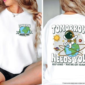 May include: White sweatshirt featuring the text "TOMORROW NEEDS YOU" and a graphic of the Earth. The back of the sweatshirt has an astronaut on the Earth with the same text and the phrase "KEEP GOING - YOU ARE NOT ALONE".