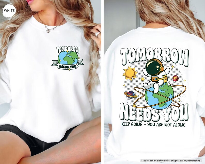 May include: White sweatshirt featuring the text "TOMORROW NEEDS YOU" and a graphic of the Earth. The back of the sweatshirt has an astronaut on the Earth with the same text and the phrase "KEEP GOING - YOU ARE NOT ALONE".