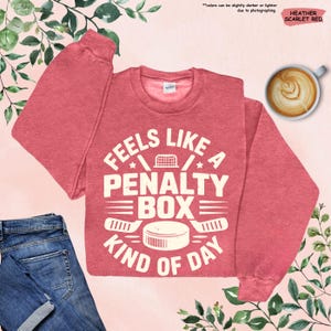 May include: Heather scarlet red sweatshirt with the text "Feels Like A Penalty Box Kind of Day" in white. The design includes hockey-related graphics such as a hockey net, hockey sticks, and a puck. A cup of coffee and a pair of jeans are also in the image.