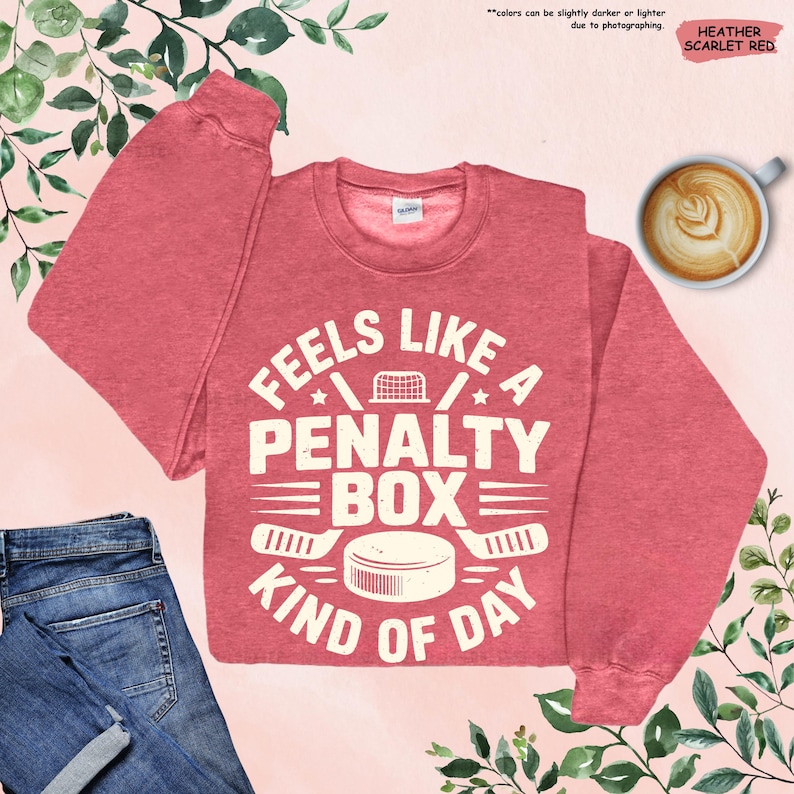 May include: Heather scarlet red sweatshirt with the text "Feels Like A Penalty Box Kind of Day" in white. The design includes hockey-related graphics such as a hockey net, hockey sticks, and a puck. A cup of coffee and a pair of jeans are also in the image.