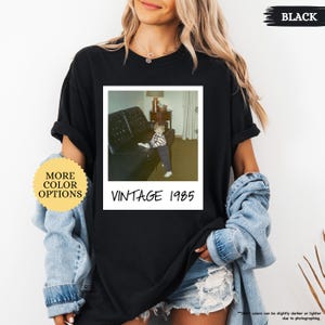 Op de afbeelding: Zwart T-shirt met een vintage foto van een jong kind dat op een bank zit. De foto is omlijst met een witte rand en de tekst "Vintage 1985" is onder de foto gedrukt.