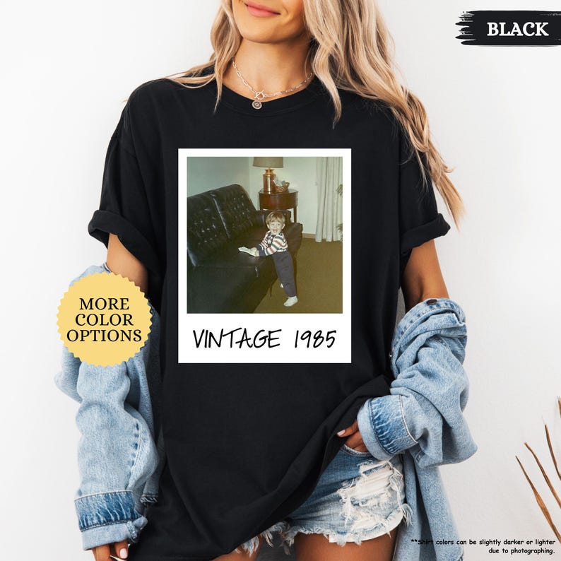 Op de afbeelding: Zwart T-shirt met een vintage foto van een jong kind dat op een bank zit. De foto is omlijst met een witte rand en de tekst "Vintage 1985" is onder de foto gedrukt.