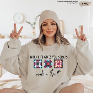 May include: Sand-colored sweatshirt with the text "WHEN LIFE GIVES YOU SCRAPS make a Quilt" and three colorful quilt block designs. The sweatshirt is a neutral color and has a crew neck.