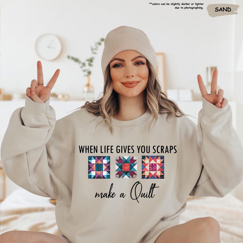 May include: Sand-colored sweatshirt with the text "WHEN LIFE GIVES YOU SCRAPS make a Quilt" and three colorful quilt block designs. The sweatshirt is a neutral color and has a crew neck.