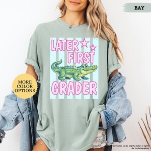 May include: A light green t-shirt with the text "LATER FIRST GRADER" in pink, along with a cartoon alligator. The shirt also has a "MORE COLOR OPTIONS" button.