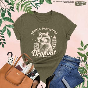 May include: Olive green t-shirt with a raccoon graphic wearing sunglasses and holding a drink. The text on the shirt reads "GENTLE PARENTING Dropout EST. THIS MORNING". The shirt is paired with blue jeans and a brown tote bag.