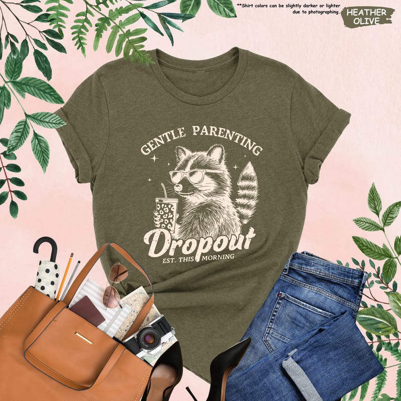 May include: Olive green t-shirt with a raccoon graphic wearing sunglasses and holding a drink. The text on the shirt reads "GENTLE PARENTING Dropout EST. THIS MORNING". The shirt is paired with blue jeans and a brown tote bag.