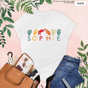 Puede incluir: Camiseta blanca con un dise&ntilde;o retro que muestra el nombre "SOPHIE" escrito en lenguaje de se&ntilde;as. Cada letra est&aacute; representada por un gesto de mano de color diferente.