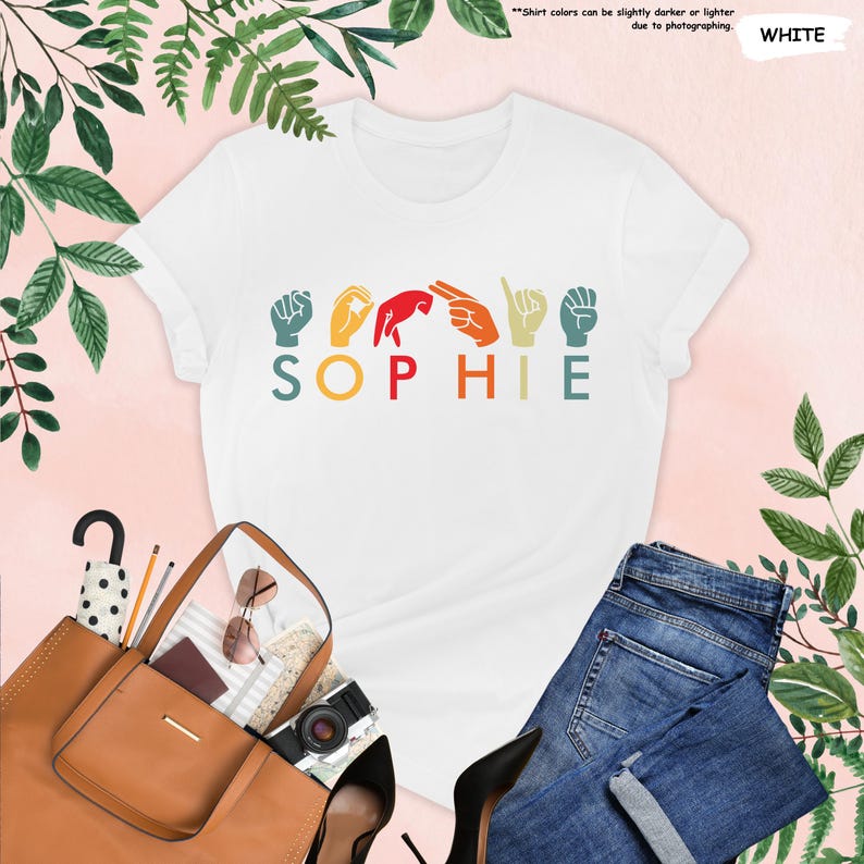 Puede incluir: Camiseta blanca con un dise&ntilde;o retro que muestra el nombre "SOPHIE" escrito en lenguaje de se&ntilde;as. Cada letra est&aacute; representada por un gesto de mano de color diferente.