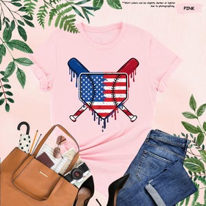 K&ouml;nnte beinhalten: Hellrosa T-Shirt mit einem Baseball-Motiv im Design der amerikanischen Flagge. Das Design umfasst gekreuzte Schl&auml;ger und tropfende Farbeffekte. Das Shirt wird mit blauen Jeans, einer braunen Tragetasche und schwarzen Abs&auml;tzen pr&auml;sentiert.