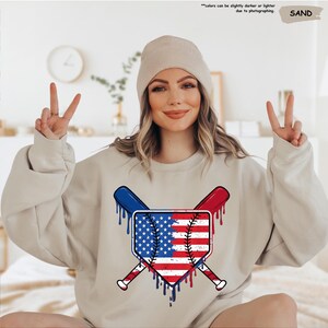 K&ouml;nnte beinhalten: Ein sandfarbenes Sweatshirt mit einem Baseball-Motiv im Design der amerikanischen Flagge. Die Grafik zeigt gekreuzte Schl&auml;ger und einen Baseball mit roten, wei&szlig;en und blauen Akzenten. Der Text oben lautet: "Farben k&ouml;nnen aufgrund der Fotografie etwas dunkler oder heller sein."