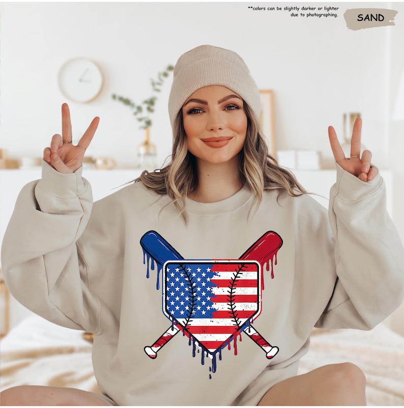 K&ouml;nnte beinhalten: Ein sandfarbenes Sweatshirt mit einem Baseball-Motiv im Design der amerikanischen Flagge. Die Grafik zeigt gekreuzte Schl&auml;ger und einen Baseball mit roten, wei&szlig;en und blauen Akzenten. Der Text oben lautet: "Farben k&ouml;nnen aufgrund der Fotografie etwas dunkler oder heller sein."
