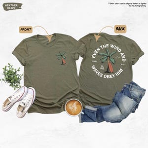 K&ouml;nnte beinhalten: Heather-olivfarbene T-Shirts mit einem Palmen- und Surfbrett-Motiv. Die Vorderseite des T-Shirts hat ein kleines Palmen-Design. Die R&uuml;ckseite des T-Shirts hat ein gr&ouml;&szlig;eres Design mit dem Text "Even the wind and waves obey him".